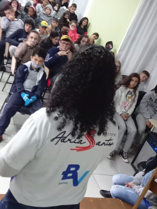 Foto de Adria de costas com camiseta do Instituto falando para crianças.